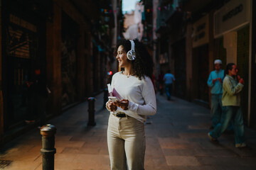 Fototapeta premium Happy young woman in white wireless headphones listening to music and holding smartphone while walking on cozy city street, cheerful female student enjoying audio podcast connection
