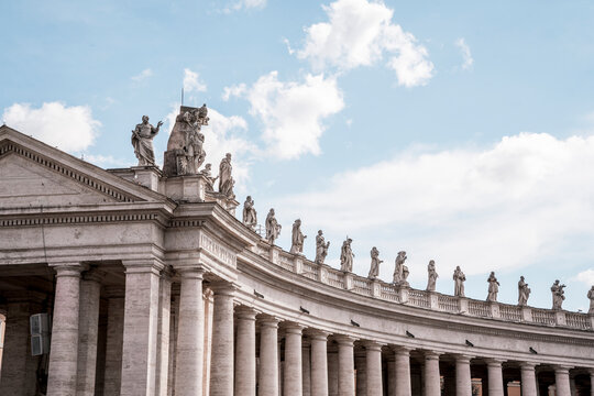 Curved classical colonnade of stone columns topped with sculpted statues under a blue sky