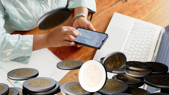 Mature woman tapping phone using finance app at desk near laptop, causing coin pile obscuring hands