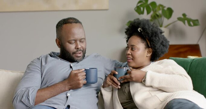 African couple sitting on couch with lav mic recording talk, woman reaching for mug, man sipping