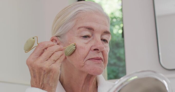 Senior woman in white robe placing green roller on cheek, rolling upward for skincare at vanity