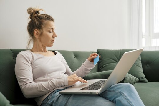 A woman engages in online shopping, holding a credit card in her hand and using her laptop on a green couch