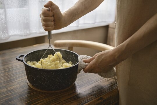 A person mashes cooked potatoes in a dark pot, using a hand masher. The cozy kitchen features natural light streaming through a window, enhancing the warm atmosphere of weekend cooking