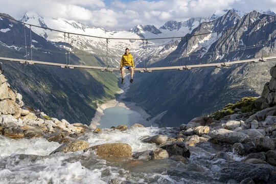 Mountaineer sitting on a suspension bridge over a mountain stream Alelebach, picturesque mountain landscape near the Olpererh&uuml;tte, view of turquoise blue lake Schlegeisspeicher, glaciated rocky mountain peaks Hoher Wei&szlig;zint and Hochfeiler with glacier Schlegeiskees, Berliner H&ouml;henweg, Zillertal Alps, Tyrol, Austria