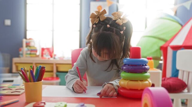 Young girl holds a pink crayon and draws on paper at a colorful playroom table with stacking toy and markers visible; concentration creativity childhood.