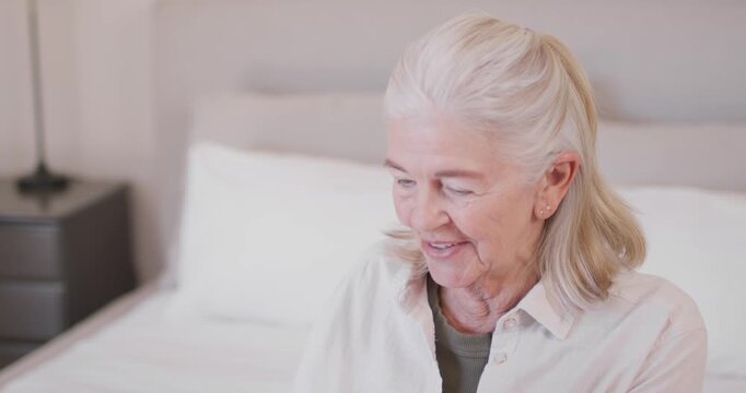Senior woman sitting in softly lit bedroom, gazing down, reaching for tablet and tapping, laughing