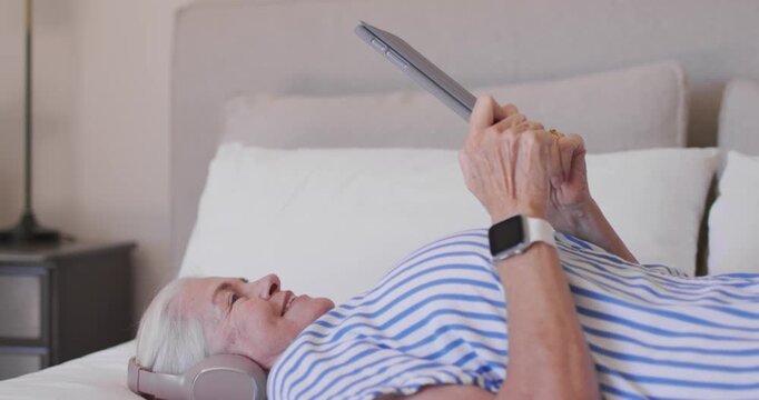 Senior female in striped shirt propped on bed wearing headset holding tablet, tilting to watch