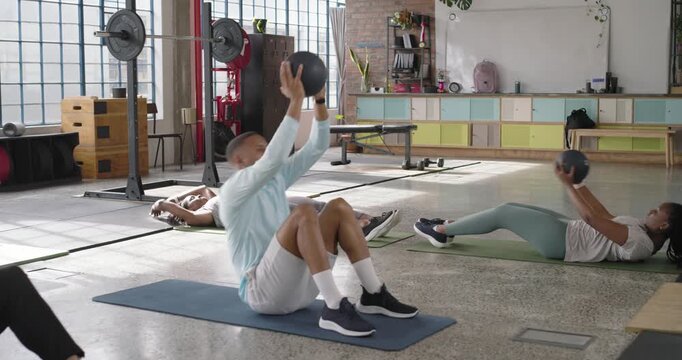Diverse group on cue doing sit-ups passing medball in gym, woman-on-mat sharing high-five finishing