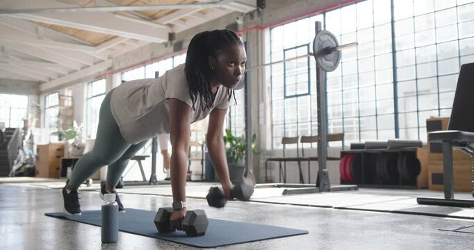African American woman holding high plank, alternating hex dumbbell rows on blue mat for core
