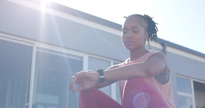 African American woman sitting on step by glass door in gym gear, sunlight causing tracker checking