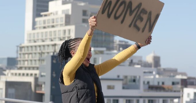African American woman in puffer vest raising WOMEN sign on rooftop, vocalizing, advocating