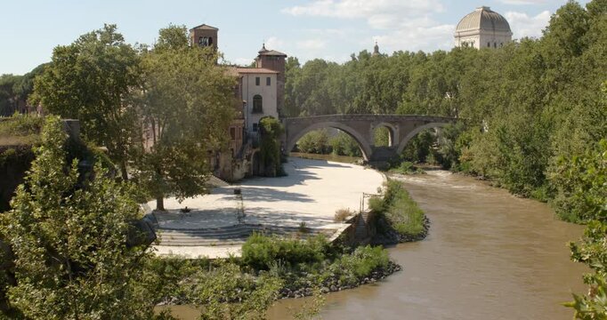 Panoramic view of Tiber Island in Rome, Italy, showcasing the ancient Pons Fabricius bridge spanning the river. Historic buildings, green trees, and the prominent synagogue dome adorn the landscape.