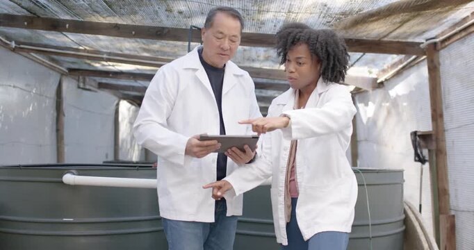 Senior Asian man and African woman in lab coats pointing at tanks tapping tablet correlating piping