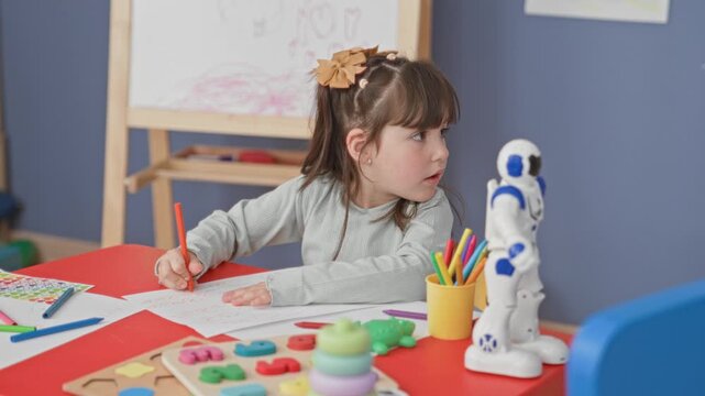 Girl holds orange pencil drawing on paper at a red table with robot toy, colored pencils, stacking rings and easel in a preschool classroom; concentration.