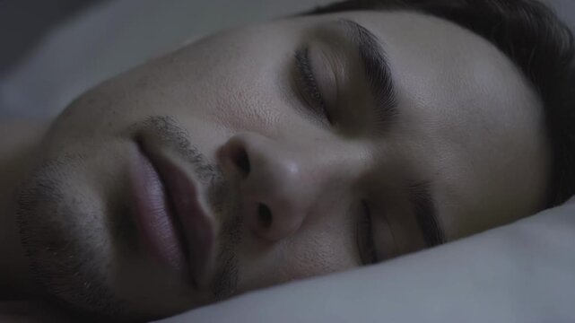 Male individual of Caucasian descent peacefully sleeping on a soft pillow, showcasing a serene expression and relaxed posture in a dimly lit bedroom environment