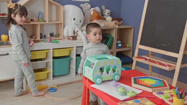 Girl holding a toy cup and reaching into a play kitchen in a bright playroom while boy stands holding a toy car; playful bonding.