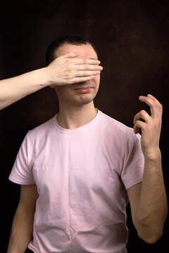Blindfolded young man making hand gesture while another hand covers eyes dramatically conceptually