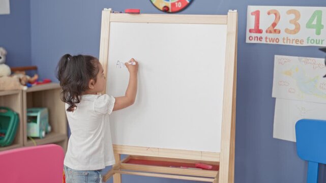 Little hispanic girl writing on easel whiteboard in classroom, marker in right hand forming letters and numbers near the top; curiosity learning play.