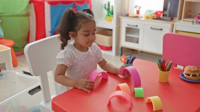 Little girl playing with colorful stacking toys, using hands to hold a pink arch on a red table in a daycare building; early learning concentration.