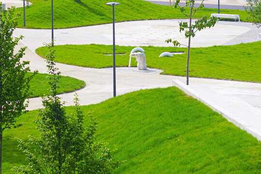 Contoured grassy landforms and intersecting pedestrian routes define newly completed public park landscape with young trees. Captures terrain-based landscape architecture and people-first placemaking.