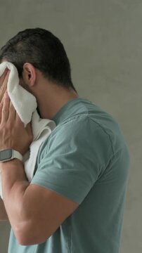 Vertical video: Reaching for white towel on wooden shelf, man in teal tee drying face at sink