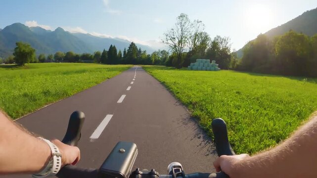 POV of cyclist riding on asphalt bike path in Slovenia. First person view of male hands on bicycle handlebar cycling on road near green alpine fields and mountains, active outdoor sport lifestyle