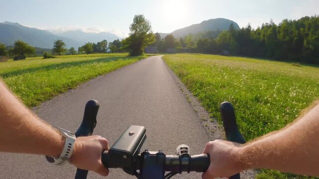POV of cyclist riding on asphalt bike path in Slovenia. First person view of male hands on bicycle handlebar cycling on road near green alpine fields and mountains, active outdoor sport lifestyle