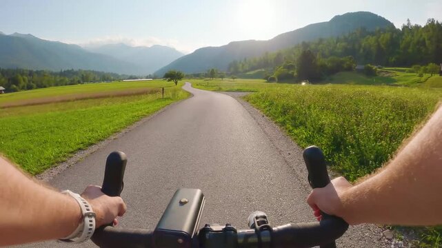 POV of cyclist riding on asphalt bike path in Slovenia. First person view of male hands on bicycle handlebar cycling on road near green alpine fields and mountains, active outdoor sport lifestyle