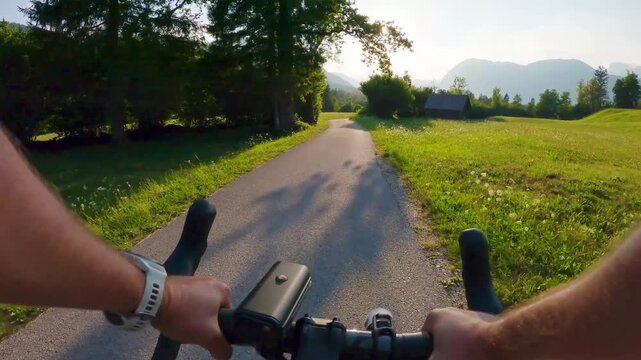 POV of cyclist riding on asphalt bike path in Slovenia. First person view of male hands on bicycle handlebar cycling on road near green alpine fields and mountains, active summer sport lifestyle