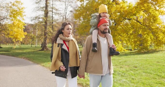 Family with mother father and daughter walking. Parents enjoy an autumn park stroll as father carries the child on shoulders, warm clothes and golden leaves. Theme of happy family togetherness.
