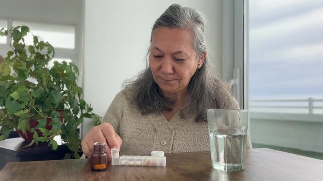 Senior woman taking daily medication while using a weekly pill organizer at home. Concept of medication management, healthcare, aging, chronic disease, treatment, retirement and healthy aging.