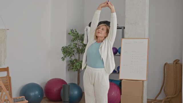Woman stretches arms overhead with interlaced fingers beside exercise balls and a workout whiteboard in a studio; calm wellbeing.