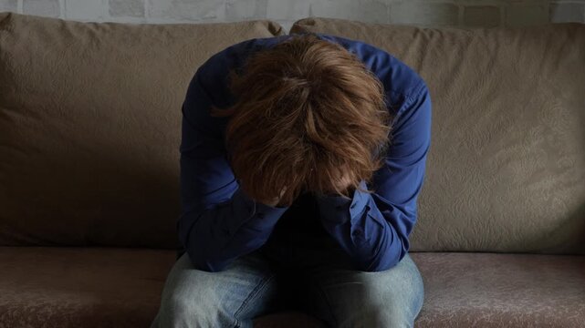 Young man (zoomer) with messy hair leaning forward
 and holding his head. Visibly troubled, feeling stress, burnout and mental exhaustion. Depression and mental health concept.