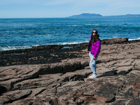 Teenager girl standing on rough stone coast, blue ocean and blue sky in the background. Travel and tourism. Mullaghmore, Ireland. Trip to amazing Irish nature. Girl in purple jacket, in dark glasses.