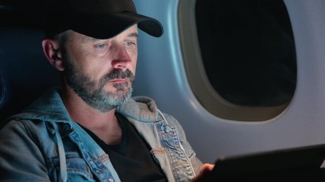 Middle-aged man with gray beard wearing black cap and denim jacket uses tablet inside airplane cabin. Screen glow illuminates his focused face near oval window during comfortable flight journey.