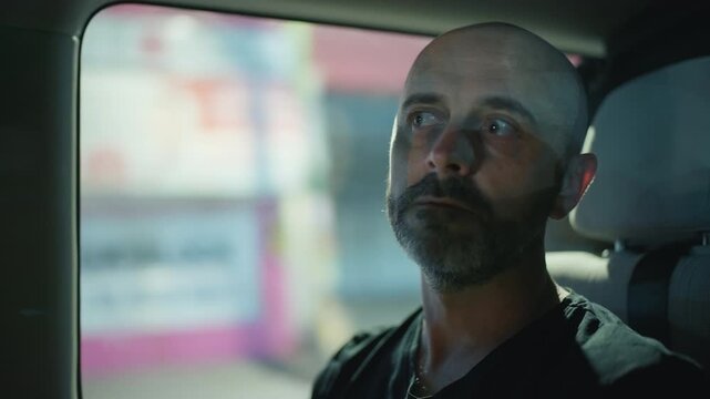 Middle-aged bald man with beard rides as passenger in minibus interior. He gazes thoughtfully through window as urban scenery passes by with colorful lights and buildings creating cinematic.