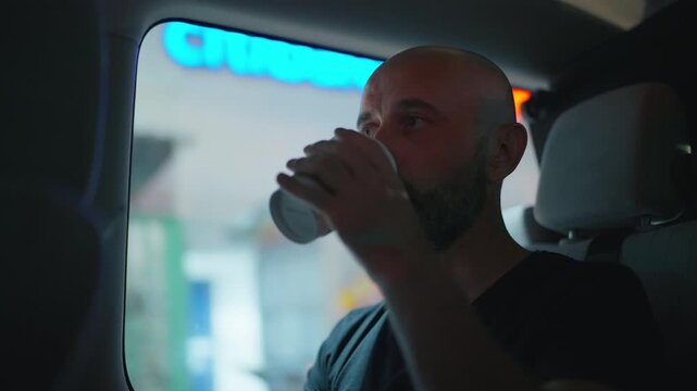 Man with beard sits in minibus interior gazing through window as urban scenery passes by. Passenger travels in shuttle van watching city streets with bokeh lights and buildings visible outside.