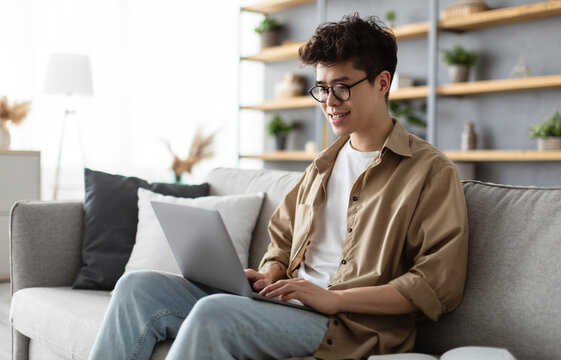 Home Office Concept. Portrait Of Smiling Asian Man In Glasses Sitting On Couch, Working On Pc Laptop Indoors In Living Room. Happy Guy Student Using Computer For Rest, Study Or Education
