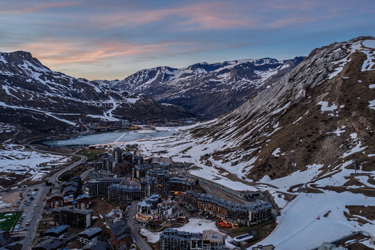 Evening drone view of Tignes 2100 in the French Alps, showing illuminated mountain resort buildings and hotels surrounded by dramatic alpine peaks under twilight sky atmosphere