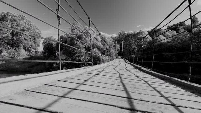 Cinematic low-angle tracking shot moving forward on a weathered wooden suspension bridge in black and white. Sunlight casts long, dramatic shadows from wire railings across the planks