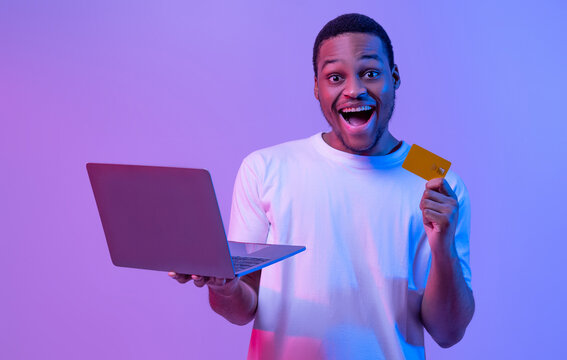 Online Shopping. Portrait of joyful black man with laptop and credit card standing in neon lighting over purple background, cheerful african guy enjoying making payments in internet, copy space