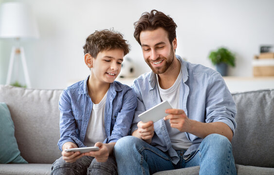 Show me your score. Boy competing with dad in online video games on smartphones, sitting on couch at home. Guy looking at father's phone, enjoying spending time together on weekend
