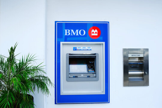 BMO ATM embedded in a white exterior wall with blue branding and red logo, front-facing cash machine and touchscreen, adjacent stainless steel night deposit slot, potted plant beside the bank facade