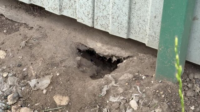 Close-up view of a corrugated metal exterior wall with green painted trim beside an uneven gravel and soil foundation. Small rocks and debris scatter