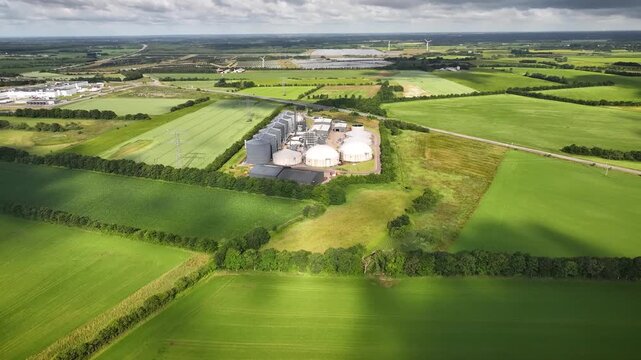 Aerial footage of a biogas plant with anaerobic digesters, revealing a mobile sprinkler irrigation system watering green agricultural fields as the drone flies backward.