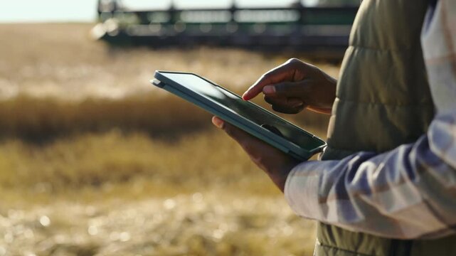 A farm worker stands in a field using a tablet to monitor crop information. The sun shines, and farm equipment is visible in the background during the day.