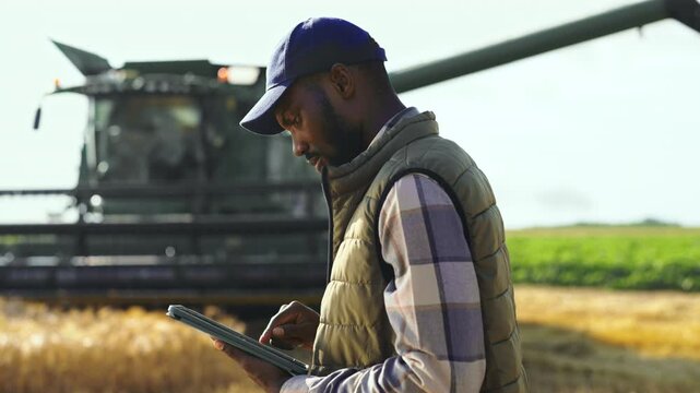 A farmer stands in a field while using a tablet to check data. In the background, a combine harvester works to gather crops. It is a sunny day on a large farm.