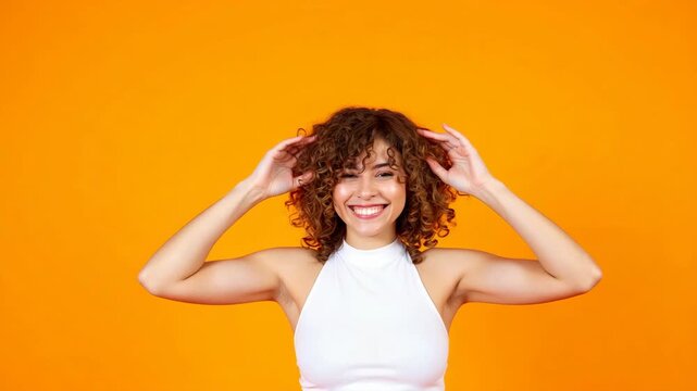 A joyful woman with curly hair and a white top is smiling broadly. She has her hands placed on her head. The background is an orange gradient which adds warmth to the .