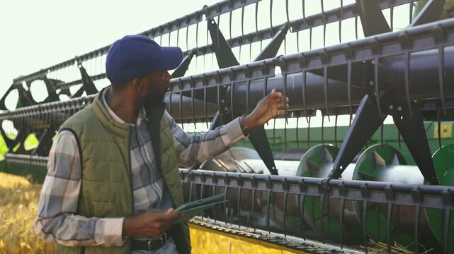 A farmer stands by a harvesting machine and uses a tablet to check data. He wears a cap and a vest. The sun shines on the field behind him while he works.