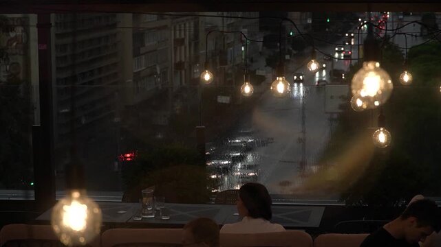 People dine at tables on the rooftop terrace of a cafe during a summer evening rain, the headlights of passing cars shining below, and loft-style interior lamps swaying in the wind from the ceiling.
